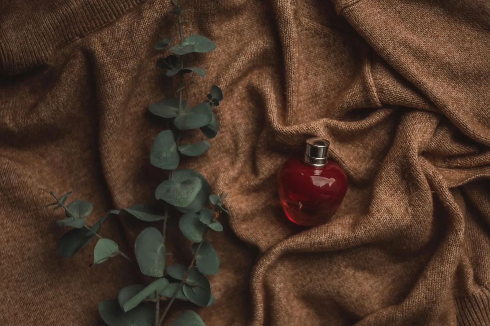 Pequeño frasco rojo de perfume sobre un tejido de punto color camello con ramas de eucalipto