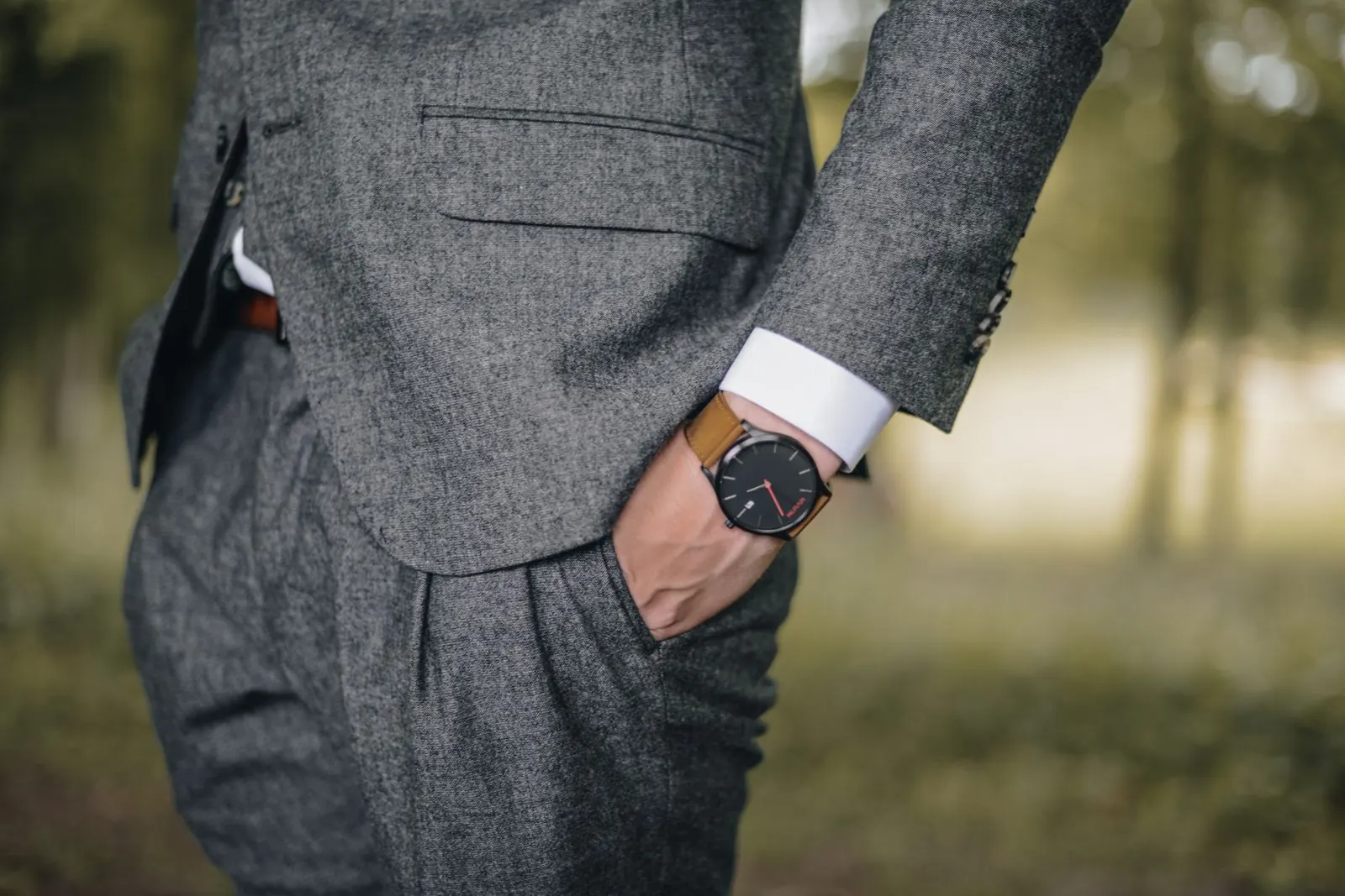 Persona con traje gris de tweed y la mano en el bolsillo, con un reloj de esfera negra y correa de cuero